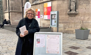 Beate Müller-Gemmeke steht vor der Citykirche neben einem Aufsteller, auf dem Informationen zu Veranstaltungen wie Kinderbasteln und Theater angezeigt werden. Sie lächelt und hält Flyer in der Hand. Im Hintergrund sind Plakate und der Eingang der Kirche zu erkennen.