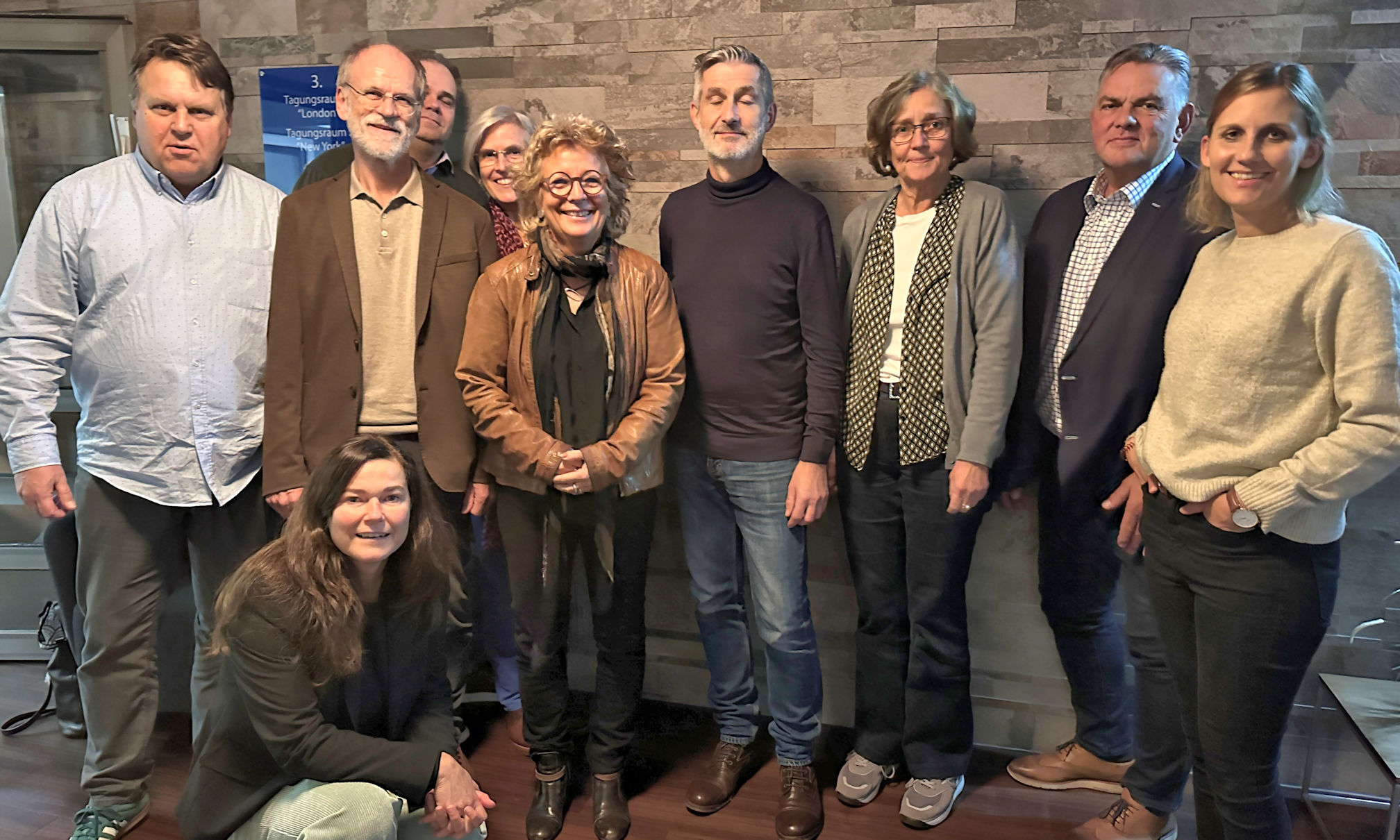 Eine Gruppe von Personen, darunter Beate Müller-Gemmeke, steht vor einer Wand mit gemusterten Fliesen. Alle lächeln in die Kamera, eine Person hockt vorne. Die Gruppe wirkt engagiert und befindet sich in einem informellen Besprechungsraum.