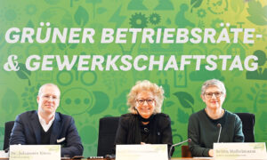 Drei Personen sitzen an einem Tisch mit Namensschildern vor einer grünen Wand mit dem Schriftzug „Grüner Betriebsräte- & Gewerkschaftstag“. Beate Müller-Gemmeke in der Mitte, links Dr. Johannes Kiess, rechts Britta Haßelmann. Darunter steht: „Demokratie verteidigen – Mitbestimmung stärken“