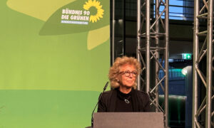 Beate Müller-Gemmeke steht an einem Rednerpult mit dem Logo von Bündnis 90/Die Grünen. Im Hintergrund ist eine grüne Wand mit dem Parteilogo und einer modernen Glasfassade zu sehen. Sie blickt konzentriert, während sie spricht.