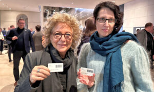 Beate Müller-Gemmeke steht mit Sabine Grützmacher vor dem Plenarsaal. Beide haben eine weiße Karte in der Hand, die sie in die Kamera halten. Im Hintergrund sind mehrere Abgeordnete