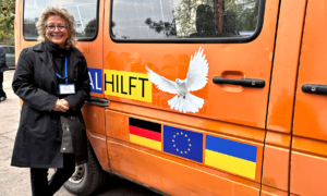 Beate Müller-Gemmeke steht vor einem orangefarbenen Van mit Beschriftung für humanitäre Hilfe. Das Fahrzeug ist mit den Flaggen der Ukraine, Deutschland und der EU und einer Friedenstaube dekoriert.