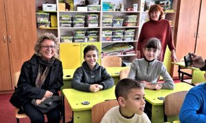 Beate Müller-Gemmeke sitzt neben drei Kindern in einem bunten Klassenzimmer mit Regalen voller Lernmaterialien. Alle lächeln in die Kamera, im Hintergrund steht eine Lehrkraft.