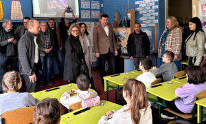 Eine Gruppe von Erwachsenen steht in einem Klassenzimmer vor einer Gruppe von Schüler:innen. Die Schüler:innen sitzen aufmerksam auf bunten Stühlen. An der Wand hängt eine Projektionsleinwand.