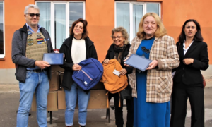 Mehrere Frauen verteilen Rucksäcke und Tablets vor einem orangefarbenen Gebäude. Eine der Frauen hält ein Tablet stolz in die Kamera. Es ist die Schulleiterin.