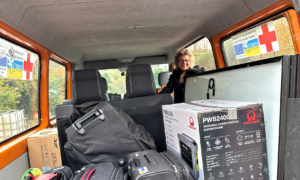 Beate Müller-Gemmeke steht in einem Transporter mit humanitärer Fracht, die für den Einsatz in einer Hilfsmission bestimmt ist. Im Vordergrund steht der Schriftzug „Solidarität heißt: hinsehen, zuhören, helfen.“ umgeben von verwischten Pinselstrichen in den Farben Blau und Gelb, die an die ukrainische Flagge erinnern. Pakete und Ausrüstungsgegenstände sind ebenfalls sichtbar. Ein Schild im Hintergrund weist auf die humanitäre Hilfe hin.