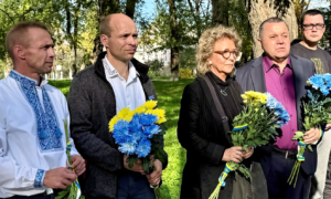 Beate Müller-Gemmeke und zwei Männer halten Sträuße in den ukrainischen Nationalfarben Blau und Gelb. Sie stehen auf einer Wiese, umgeben von Bäumen, in einer feierlichen, sehr ernsten Atmosphäre.