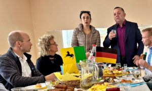 Beate Müller-Gemmeke und andere Personen sitzen an einem gedeckten Tisch mit verschiedenen Speisen. Im Hintergrund sind deutsche und ukrainische Flaggen sichtbar, während der Bürgermeister eine Rede hält.