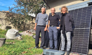 Beate Müller-Gemmeke und zwei Männer stehen vor einer Solaranlage in einem ländlichen Gebiet. Im Hintergrund ist ein kleineres Gebäude mit Dach und Bäumen zu sehen.