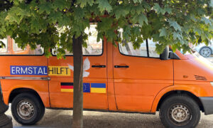 Ein orangefarbenes Transportfahrzeug mit der Aufschrift „Ermstal hilft“, verziert mit einer weißen Friedenstaube sowie den Flaggen Deutschlands, der EU und der Ukraine. Es steht unter einem Baum, der seine Blätter über das Fahrzeug ausbreitet.