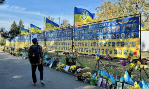 Eine Gedenkallee in Mykolajiw, die mit Bildern von Gefallenen geschmückt ist. Die Tafeln mit Porträts sind von Blumen und ukrainischen Flaggen umgeben. Ein Mann mit Rucksack läuft entlang der Gedenktafeln.