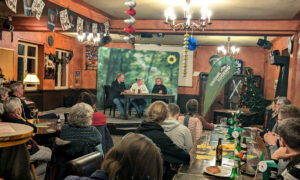 Eine Podiumsdiskussion in einem Veranstaltungsraum. Drei Personen sitzen auf der Bühne an einem Tisch, während das Publikum im Vordergrund zuhört. Auf der Bühne sind ein Mann, eine Frau im weißen Pullover und Beate Müller-Gemmeke. Im Hintergrund ein grünes Banner mit Sonnenblumenlogo.