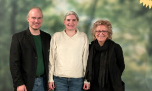 Gruppenfoto vor einem Hintergrund mit Waldmotiv. Drei Personen stehen nebeneinander: links ein Mann, in der Mitte Susanne Floss im weißen Pullover und rechts Beate Müller-Gemmeke, alle lächeln in die Kamera.