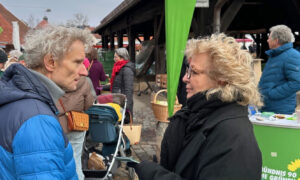 Eine Frau mit lockigem, blondem Haar spricht mit einem Mann mit grauem Haar auf einem öffentlichen Platz. Es ist Beate Müller-Gemmeke. Sie trägt eine schwarze Jacke und Handschuhe, während der Mann eine blaue Steppjacke trägt. Im Hintergrund sind ein Stand mit grünem Banner und weitere Menschen zu sehen.