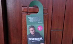 Ein Türhänger mit grünem Hintergrund ist an einer Holztür angebracht. Darauf ist ein Foto von Robert Habeck sowie der Schriftzug „ZUVER SICHT. Ein Mensch. Ein Wort.“ zu sehen. Es gibt einen Hinweis auf den Wahltermin am 23. Februar, begleitet von einer kurzen Botschaft.