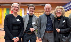 Vier Personen posieren in einer Turnhalle für ein Gruppenfoto. Beate Müller-Gemmeke steht ganz rechts, neben ihr ein älterer Herr im grauen Anzug. Links davon eine Frau in schwarzer Kleidung und ein junger Mann mit grauer Jacke. Die Stimmung ist herzlich.