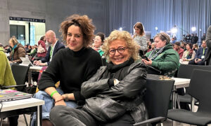 Beate Müller-Gemmeke sitzt neben einer Kollegin in einem Tagungsraum. Beide lachen herzlich in die Kamera. Im Hintergrund sind weitere Delegierte an Tischen bei der Arbeit.