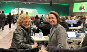 Beate Müller-Gemmeke und eine Kollegin sitzen an einem Tisch in einem Tagungsraum. Beide lächeln in die Kamera, während im Hintergrund Delegierte und Bildschirme mit Konferenzinhalten zu sehen sind.