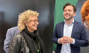 Beate Müller-Gemmeke unterhält sich lachend mit dem Bundesvorsitzenden Felix Banaszak. Beide stehen vor einer grünen Parteifahne mit Sonnenblumen-Logo, während sich im Hintergrund weitere Teilnehmer*innen befinden.