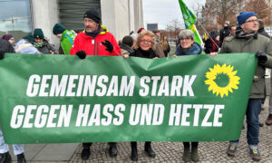 Drei Menschen halten ein großes grünes Banner mit der Aufschrift „GEMEINSAM STARK GEGEN HASS UND HETZE“. Beate Müller-Gemmeke ist eine dieser Personen. Im Hintergrund sind weitere Demonstrierende mit Grünen-Fahnen zu sehen.