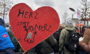 Ein rotes Pappherz mit der handgeschriebenen Aufschrift „HERZ STATT MERZ“ und einer kleinen, verzierten Figur darauf. Dahinter eine Menschenmenge bei der Demo.