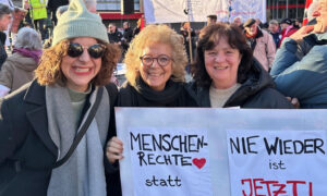 Beate Müller-Gemmeke steht mit zwei Frauen auf einer Demonstration. Sie halten zwei Schilder: „Menschenrechte statt rechte Menschen“ mit einem roten Herz und „Nie wieder ist jetzt! Demokratie ist nicht verhandelbar.“ Im Hintergrund sind viele Menschen, eine Bühne und ein Banner mit der Aufschrift „Kampf dem Faschismus“ zu sehen.