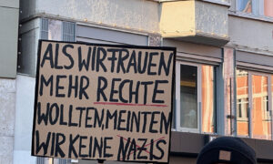 Ein großes, handgeschriebenes Plakat wird hochgehalten. Darauf steht in großen Buchstaben: „Als wir Frauen mehr Rechte wollten, meinten wir keine Nazis.“ Mehrere Wörter sind rot unterstrichen. Im Hintergrund sind Gebäude mit Fenstern und ein Balkon zu erkennen.