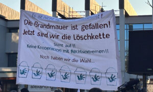 Ein großes Transparent wird auf einer Demonstration gehalten. Darauf steht: „Die Brandmauer ist gefallen! Jetzt sind wir die Löschkette.“ Unten sind gezeichnete Wassereimer mit Handabdrücken. Weitere Schriftzüge fordern, sich gegen Rechtsextremismus zu stellen. Im Hintergrund sind Gebäude zu sehen.