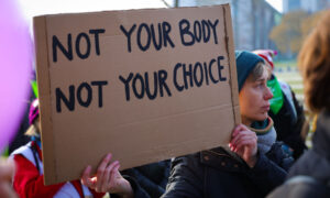 Ein Pappschild mit der englischen Aufschrift „NOT YOUR BODY, NOT YOUR CHOICE“ wird von einer Person hochgehalten. Der Hintergrund ist leicht verschwommen, es ist eine Protestszene mit weiteren Menschen zu erahnen.