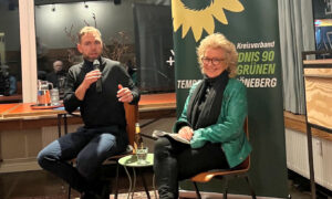 Beate Müller-Gemmeke und Moritz Heuberger sitzen auf der Bühne einer Podiumsdiskussion. Moritz Heuberger spricht mit einem Mikrofon in der Hand, während Beate Müller-Gemmeke lächelnd zuhört und Notizen hält. Hinter ihnen steht ein grünes Banner mit dem Logo von Bündnis 90/Die Grünen und der Aufschrift „Kreisverband Tempelhof-Schöneberg“.