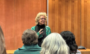 Beate Müller-Gemmeke steht vor einer hölzernen Wand und spricht zu einer Gruppe von Zuhörer:innen. Sie trägt eine grüne Lederjacke und eine schwarze Brille. Die Zuhörer:innen sitzen mit dem Rücken zur Kamera, während sie ihr aufmerksam folgen.