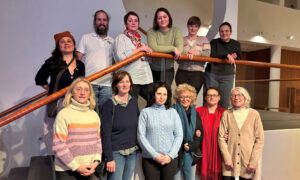 Eine Gruppe von zwölf Personen, darunter Beate Müller-Gemmeke, posiert auf einer Treppe im Bundestag. Die Teilnehmenden, größtenteils Frauen, stehen und sitzen entspannt zusammen. Sie tragen winterliche Kleidung und wirken engagiert und interessiert.