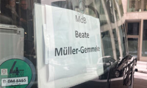 Nahaufnahme eines Schildes hinter der Windschutzscheibe eines Busses. Darauf steht „MdB Beate Müller-Gemmeke“. Im Spiegelbild der Scheibe sind Personen im Bus zu erkennen. Draußen ist es winterlich mit leichtem Schneefall.