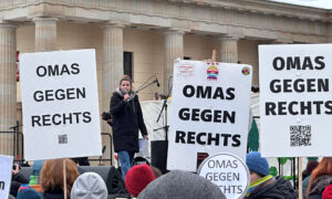 Eine junge Frau spricht mit einem Mikrofon auf einer Bühne während einer Demonstration vor dem Brandenburger Tor. Im Vordergrund halten Menschen Schilder mit der Aufschrift „Omas gegen Rechts“ hoch.