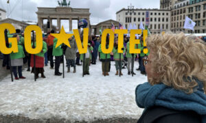 Beate Müller-Gemmeke beobachtet eine Gruppe Demonstrierender, die große gelbe Buchstaben in die Höhe halten und das Wort „VOTE!“ formen. Der Boden ist mit Schnee bedeckt, im Hintergrund das Brandenburger Tor.