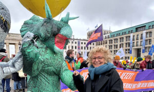 Beate Müller-Gemmeke steht lachend neben einer Person in einem großen grünen Dinosaurier-Kostüm. Im Hintergrund sind weitere Demonstrierende mit Fahnen und Bannern zu sehen.