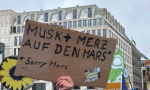 Ein selbstgemachtes Schild aus Pappe wird hochgehalten. Darauf steht: „Musk + Merz auf den Mars – Sorry Mars.“ Im Hintergrund weitere Demonstrierende mit Plakaten und Fahnen.