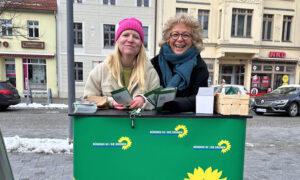 Beate Müller-Gemmeke und Jenifer Howel stehen an einem grünen Infostand von Bündnis 90/Die Grünen in Finsterwalde. Beide halten Flyer in den Händen und lächeln in die Kamera. Der Infostand ist mit dem Logo der Grünen versehen. Im Hintergrund sind Altbauten und ein schneebedeckter Gehweg zu sehen.