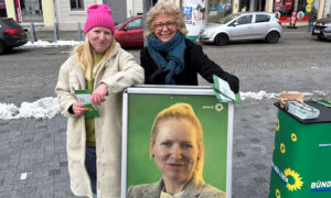 Beate Müller-Gemmeke und Jenifer Howel stehen in Finsterwalde neben einem Plakat mit Howels Porträt und dem Slogan „Entschlossenheit – Jenifer Howel für Brandenburg“. Beide halten Flyer in den Händen und lächeln. Die Straße ist mit Schnee bedeckt, im Hintergrund sind Geschäfte und Autos zu sehen.