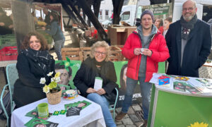 Beate Müller-Gemmeke sitzt an einem kleinen Tisch mit Flyern, einer grünen Tasse und einem Blumentopf bei einem Infostand von Bündnis 90/Die Grünen. Neben ihr sitzt eine junge Frau mit dunklen Locken. Auf der anderen stehen zwei weitere Personen: ein Mann mit roter Jacke und grauer Mütze sowie ein weiterer Mann mit Brille und Bart. Hinter ihnen sind Wahlplakate, ein Marktstand und eine hölzerne Überdachung zu sehen.