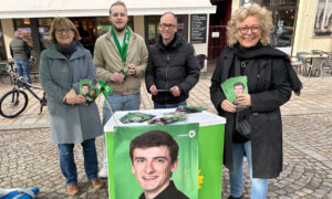 Beate Müller-Gemmeke steht mit drei weiteren Personen an einem Infostand von Bündnis 90/Die Grünen. Neben ihr eine Frau mit Brille und grauem Mantel, ein junger Mann mit grünem Schal und ein Mann mit schwarzer Jacke. Auf dem Tisch steht ein großes Wahlplakat mit dem Porträt von Jaron Immer. Alle halten Flyer in der Hand, im Hintergrund ist eine Fußgängerzone mit Gebäuden zu sehen.