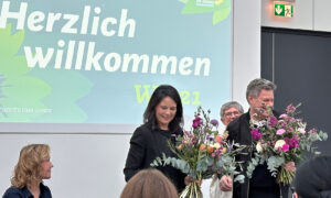 Annalena Baerbock und Robert Habeck stehen in einem großen Raum vor einer Leinwand mit der Aufschrift „Herzlich Willkommen“. Beide halten große Blumensträuße und lächeln. Im Hintergrund ist das Logo von Bündnis 90/Die Grünen zu sehen. Die Szene wirkt feierlich und wertschätzend.