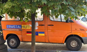 Ein orangefarbener Bus mit der Aufschrift „Ermstal hilft“ und den Flaggen Deutschlands, der EU und der Ukraine steht unter einem Baum. Eine Friedenstaube ist auf dem Bus abgebildet.