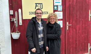 Zwei Personen stehen lächelnd vor einem roten Gebäude mit einem Schild „Möbelkammer FAU Sondershausen“. Es ist Beate Müller-Gemmeke und Kai Klemm-Lorenz. Beide tragen warme Kleidung. Die Atmosphäre wirkt freundlich und herzlich.