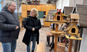 Ein Mann und Beate Müller-Gemmeke stehen in einer Werkstatt und betrachten gemeinsam ein liebevoll gestaltetes Holz-Vogelhaus mit einem „Willkommen“-Schild. Im Hintergrund sind weitere Holzarbeiten und Werkzeuge zu sehen.