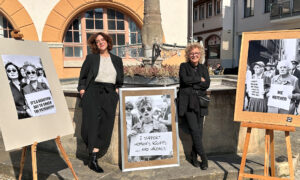 Beate Müller-Gemmeke und eine weitere Frau stehen neben drei großformatigen schwarz-weißen Protestbildern mit feministischen Botschaften. Die Plakate zeigen Frauen mit Schildern wie „It’s a beautiful day to smash the patriarchy“ und „I support women’s rights … and wrongs“. Sie lehnen sich entspannt an eine Steinmauer, im Hintergrund ein historisches Gebäude mit orangefarbenen Akzenten.