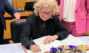 Beate Müller-Gemmeke sitzt an einem Tisch und trägt sich mit einem Füller in das Goldene Buch der Stadt ein. Muhterem Aras, in pinkem Hosenanzug, steht daneben und schaut ihr lächelnd über die Schulter. Der Tisch ist mit einem weißen Tuch und einem Blumenarrangement dekoriert.